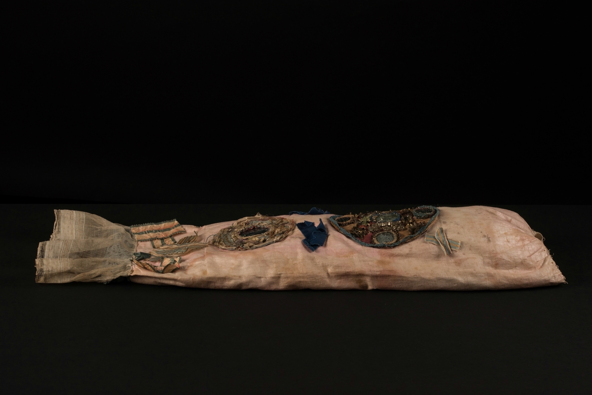 Doppåse till nyfödd. Utsidan sydd av rosarött linnetyg, mycket blekt. Påsen avsmalnande upptill med lagda veck och sprund i ryggen. Har troligtvis burits med en krage eller krås. 

Påsens nedre del avslutas med en blå spets under tyll i vitt med blå inslag. 

Påsen är dekorerad med sidenband och bomullsband som bildar kryss samt en blå rosett. 

På påsens mittfält sitter två runda dekorationer den ena 150 mm i diameter och den andra 120 mm i diameter. Den större dekorationen har 5 speglar och metalltrådar som bildar blommor. Runt hela sitter en blå spets. Den mindre dekorationen har en större spegel, denband och blommor i metalltråd.