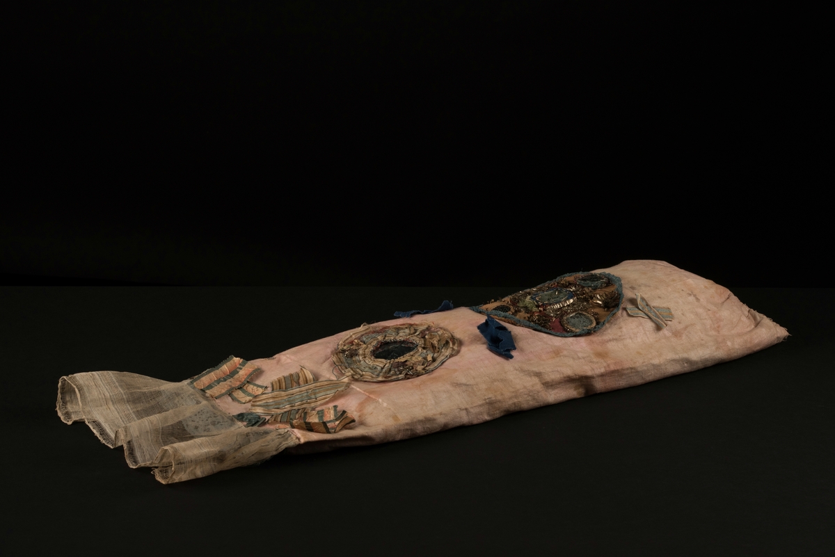 Doppåse till nyfödd. Utsidan sydd av rosarött linnetyg, mycket blekt. Påsen avsmalnande upptill med lagda veck och sprund i ryggen. Har troligtvis burits med en krage eller krås. 

Påsens nedre del avslutas med en blå spets under tyll i vitt med blå inslag. 

Påsen är dekorerad med sidenband och bomullsband som bildar kryss samt en blå rosett. 

På påsens mittfält sitter två runda dekorationer den ena 150 mm i diameter och den andra 120 mm i diameter. Den större dekorationen har 5 speglar och metalltrådar som bildar blommor. Runt hela sitter en blå spets. Den mindre dekorationen har en större spegel, denband och blommor i metalltråd.