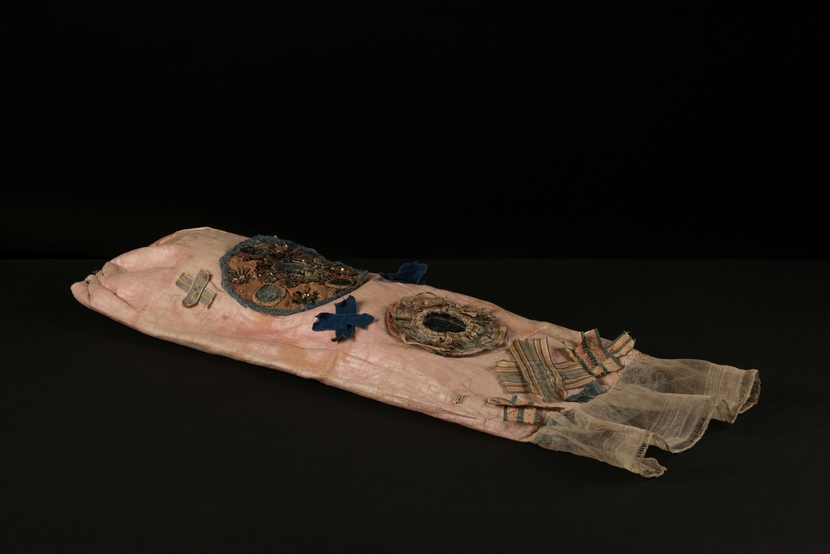 Doppåse till nyfödd. Utsidan sydd av rosarött linnetyg, mycket blekt. Påsen avsmalnande upptill med lagda veck och sprund i ryggen. Har troligtvis burits med en krage eller krås. 

Påsens nedre del avslutas med en blå spets under tyll i vitt med blå inslag. 

Påsen är dekorerad med sidenband och bomullsband som bildar kryss samt en blå rosett. 

På påsens mittfält sitter två runda dekorationer den ena 150 mm i diameter och den andra 120 mm i diameter. Den större dekorationen har 5 speglar och metalltrådar som bildar blommor. Runt hela sitter en blå spets. Den mindre dekorationen har en större spegel, denband och blommor i metalltråd.