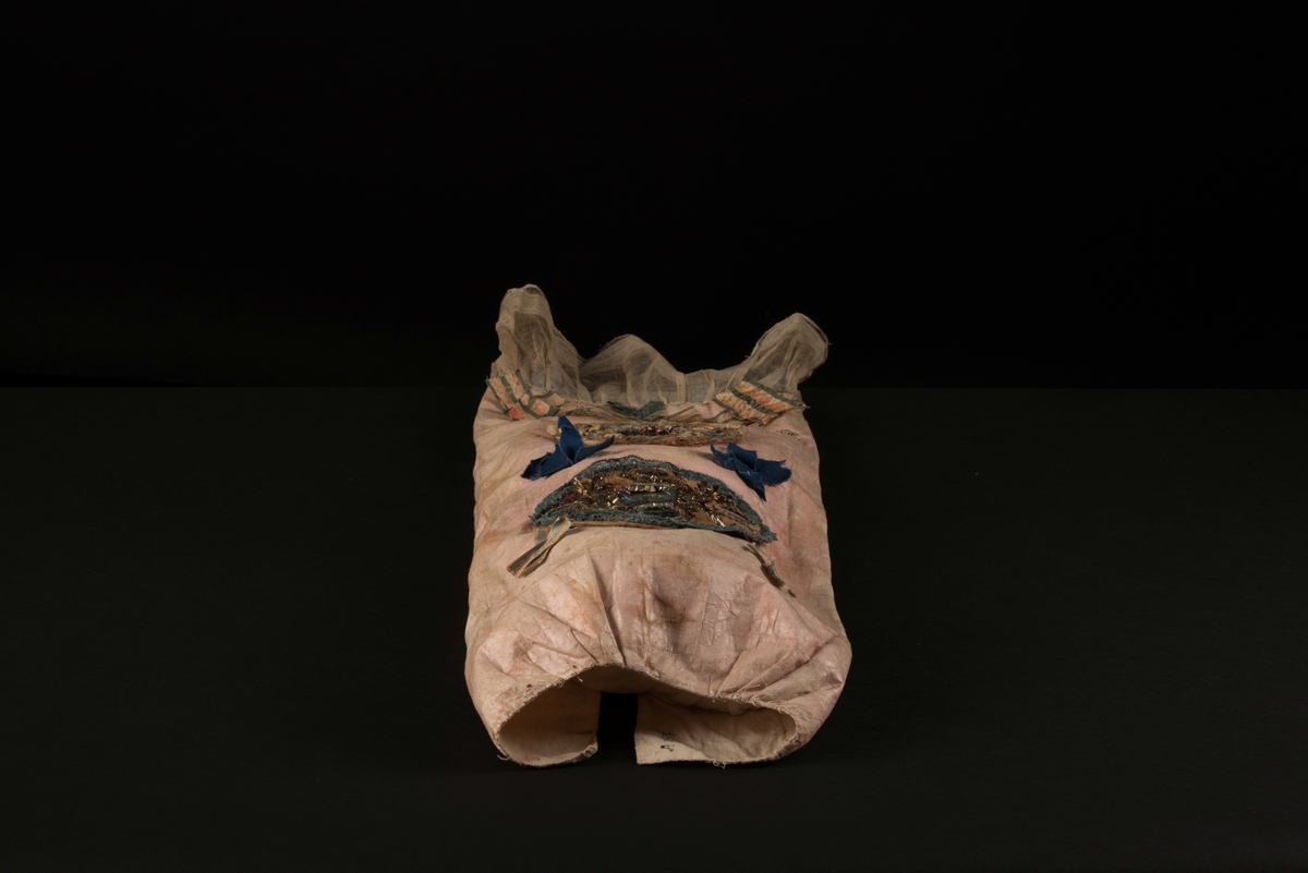 Doppåse till nyfödd. Utsidan sydd av rosarött linnetyg, mycket blekt. Påsen avsmalnande upptill med lagda veck och sprund i ryggen. Har troligtvis burits med en krage eller krås. 

Påsens nedre del avslutas med en blå spets under tyll i vitt med blå inslag. 

Påsen är dekorerad med sidenband och bomullsband som bildar kryss samt en blå rosett. 

På påsens mittfält sitter två runda dekorationer den ena 150 mm i diameter och den andra 120 mm i diameter. Den större dekorationen har 5 speglar och metalltrådar som bildar blommor. Runt hela sitter en blå spets. Den mindre dekorationen har en större spegel, denband och blommor i metalltråd.