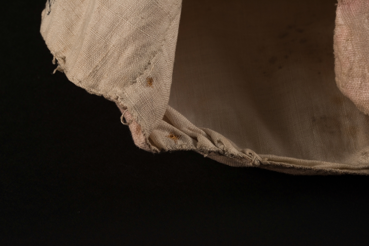 Doppåse till nyfödd. Utsidan sydd av rosarött linnetyg, mycket blekt. Påsen avsmalnande upptill med lagda veck och sprund i ryggen. Har troligtvis burits med en krage eller krås. 

Påsens nedre del avslutas med en blå spets under tyll i vitt med blå inslag. 

Påsen är dekorerad med sidenband och bomullsband som bildar kryss samt en blå rosett. 

På påsens mittfält sitter två runda dekorationer den ena 150 mm i diameter och den andra 120 mm i diameter. Den större dekorationen har 5 speglar och metalltrådar som bildar blommor. Runt hela sitter en blå spets. Den mindre dekorationen har en större spegel, denband och blommor i metalltråd.