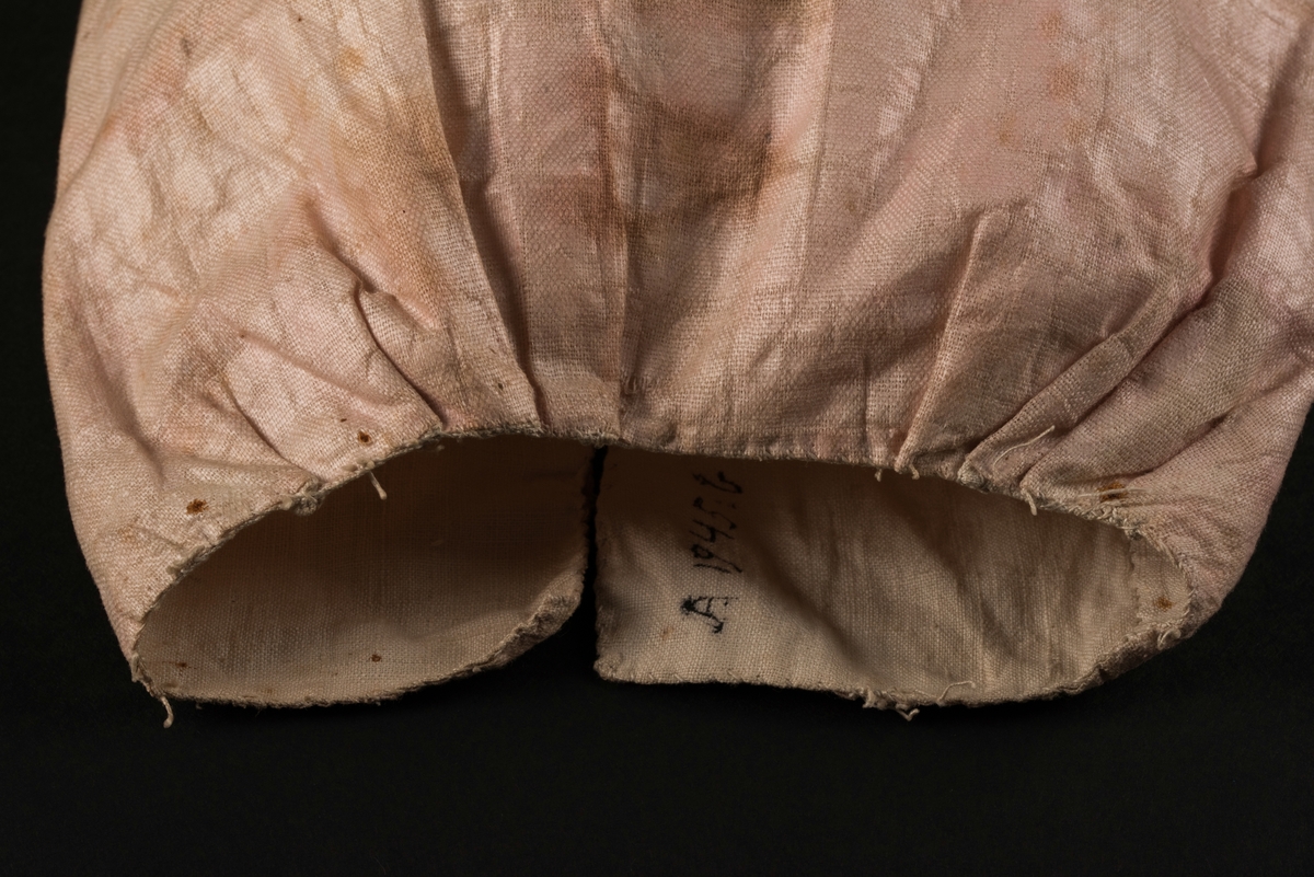 Doppåse till nyfödd. Utsidan sydd av rosarött linnetyg, mycket blekt. Påsen avsmalnande upptill med lagda veck och sprund i ryggen. Har troligtvis burits med en krage eller krås. 

Påsens nedre del avslutas med en blå spets under tyll i vitt med blå inslag. 

Påsen är dekorerad med sidenband och bomullsband som bildar kryss samt en blå rosett. 

På påsens mittfält sitter två runda dekorationer den ena 150 mm i diameter och den andra 120 mm i diameter. Den större dekorationen har 5 speglar och metalltrådar som bildar blommor. Runt hela sitter en blå spets. Den mindre dekorationen har en större spegel, denband och blommor i metalltråd.