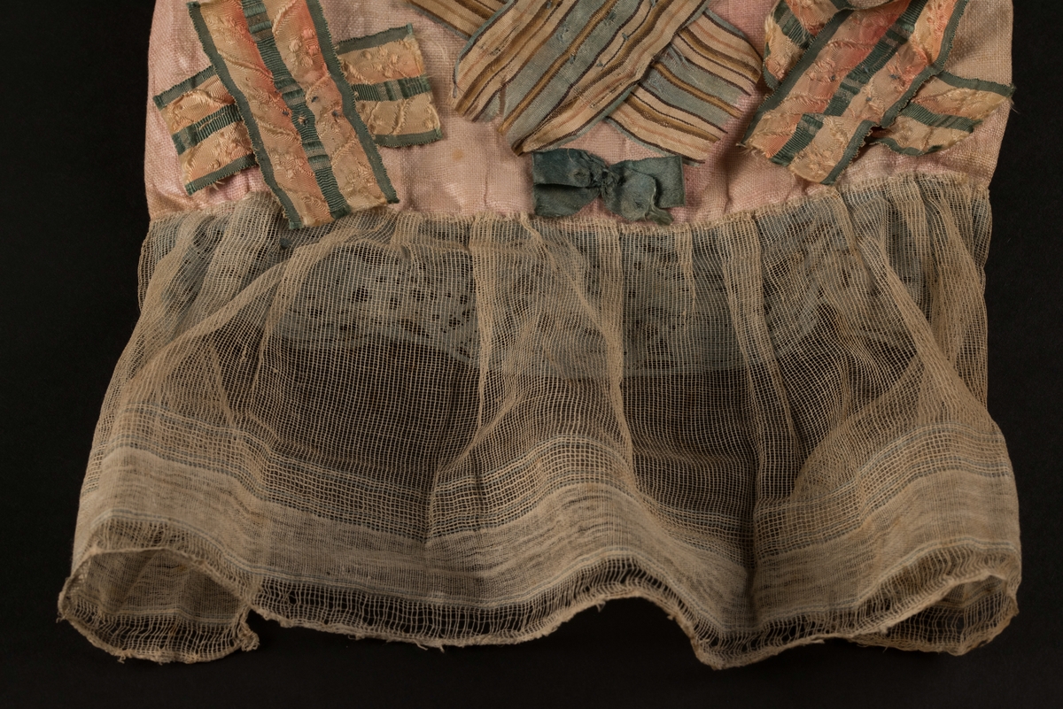 Doppåse till nyfödd. Utsidan sydd av rosarött linnetyg, mycket blekt. Påsen avsmalnande upptill med lagda veck och sprund i ryggen. Har troligtvis burits med en krage eller krås. 

Påsens nedre del avslutas med en blå spets under tyll i vitt med blå inslag. 

Påsen är dekorerad med sidenband och bomullsband som bildar kryss samt en blå rosett. 

På påsens mittfält sitter två runda dekorationer den ena 150 mm i diameter och den andra 120 mm i diameter. Den större dekorationen har 5 speglar och metalltrådar som bildar blommor. Runt hela sitter en blå spets. Den mindre dekorationen har en större spegel, denband och blommor i metalltråd.