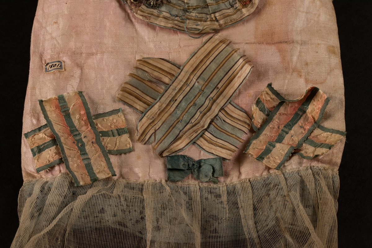 Doppåse till nyfödd. Utsidan sydd av rosarött linnetyg, mycket blekt. Påsen avsmalnande upptill med lagda veck och sprund i ryggen. Har troligtvis burits med en krage eller krås. 

Påsens nedre del avslutas med en blå spets under tyll i vitt med blå inslag. 

Påsen är dekorerad med sidenband och bomullsband som bildar kryss samt en blå rosett. 

På påsens mittfält sitter två runda dekorationer den ena 150 mm i diameter och den andra 120 mm i diameter. Den större dekorationen har 5 speglar och metalltrådar som bildar blommor. Runt hela sitter en blå spets. Den mindre dekorationen har en större spegel, denband och blommor i metalltråd.