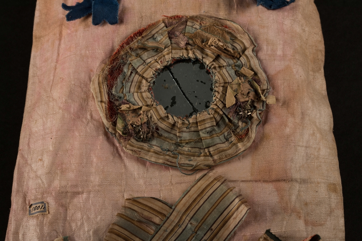 Doppåse till nyfödd. Utsidan sydd av rosarött linnetyg, mycket blekt. Påsen avsmalnande upptill med lagda veck och sprund i ryggen. Har troligtvis burits med en krage eller krås. 

Påsens nedre del avslutas med en blå spets under tyll i vitt med blå inslag. 

Påsen är dekorerad med sidenband och bomullsband som bildar kryss samt en blå rosett. 

På påsens mittfält sitter två runda dekorationer den ena 150 mm i diameter och den andra 120 mm i diameter. Den större dekorationen har 5 speglar och metalltrådar som bildar blommor. Runt hela sitter en blå spets. Den mindre dekorationen har en större spegel, denband och blommor i metalltråd.