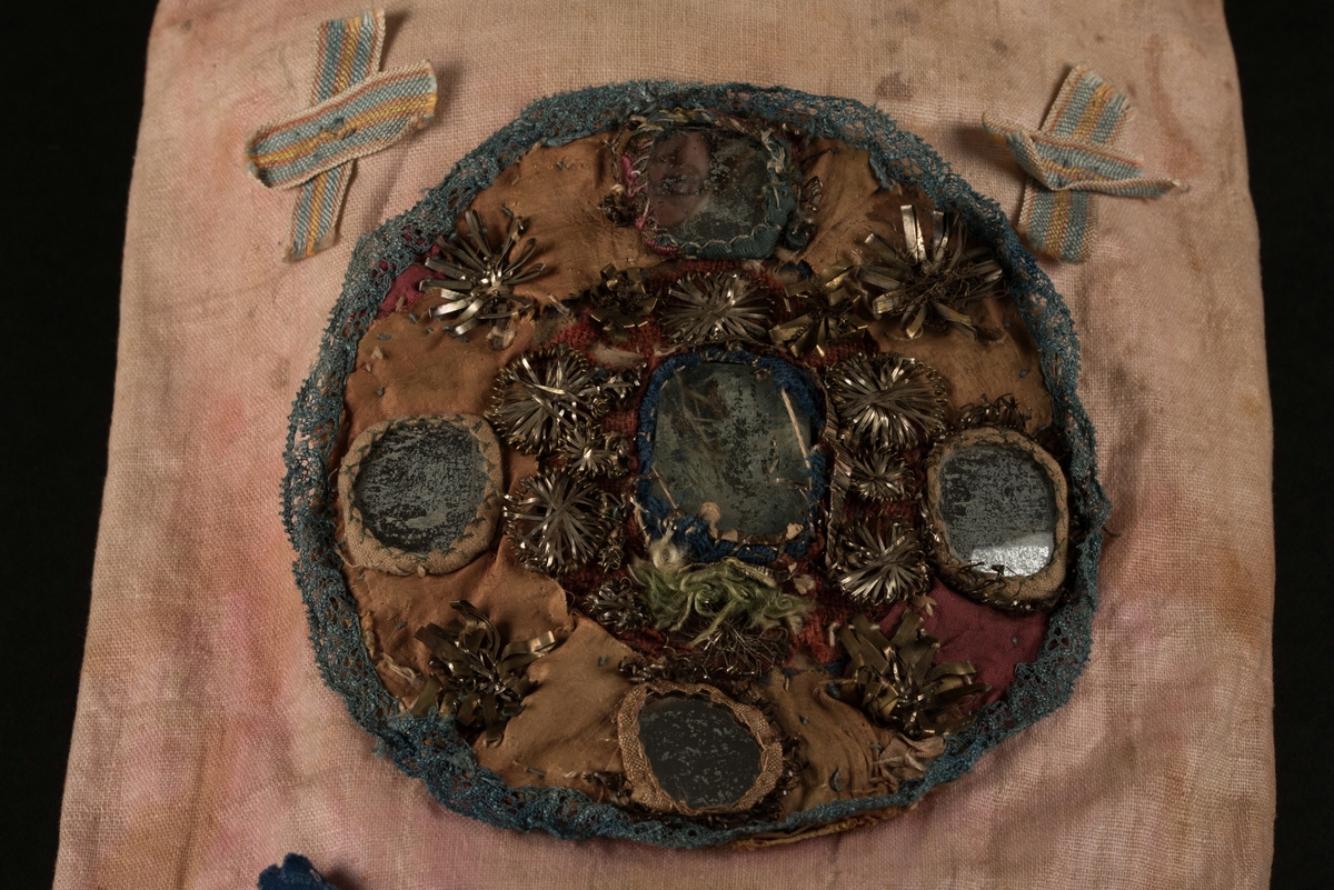 Doppåse till nyfödd. Utsidan sydd av rosarött linnetyg, mycket blekt. Påsen avsmalnande upptill med lagda veck och sprund i ryggen. Har troligtvis burits med en krage eller krås. 

Påsens nedre del avslutas med en blå spets under tyll i vitt med blå inslag. 

Påsen är dekorerad med sidenband och bomullsband som bildar kryss samt en blå rosett. 

På påsens mittfält sitter två runda dekorationer den ena 150 mm i diameter och den andra 120 mm i diameter. Den större dekorationen har 5 speglar och metalltrådar som bildar blommor. Runt hela sitter en blå spets. Den mindre dekorationen har en större spegel, denband och blommor i metalltråd.