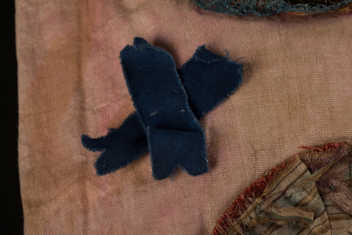 Doppåse till nyfödd. Utsidan sydd av rosarött linnetyg, mycket blekt. Påsen avsmalnande upptill med lagda veck och sprund i ryggen. Har troligtvis burits med en krage eller krås. 

Påsens nedre del avslutas med en blå spets under tyll i vitt med blå inslag. 

Påsen är dekorerad med sidenband och bomullsband som bildar kryss samt en blå rosett. 

På påsens mittfält sitter två runda dekorationer den ena 150 mm i diameter och den andra 120 mm i diameter. Den större dekorationen har 5 speglar och metalltrådar som bildar blommor. Runt hela sitter en blå spets. Den mindre dekorationen har en större spegel, denband och blommor i metalltråd.