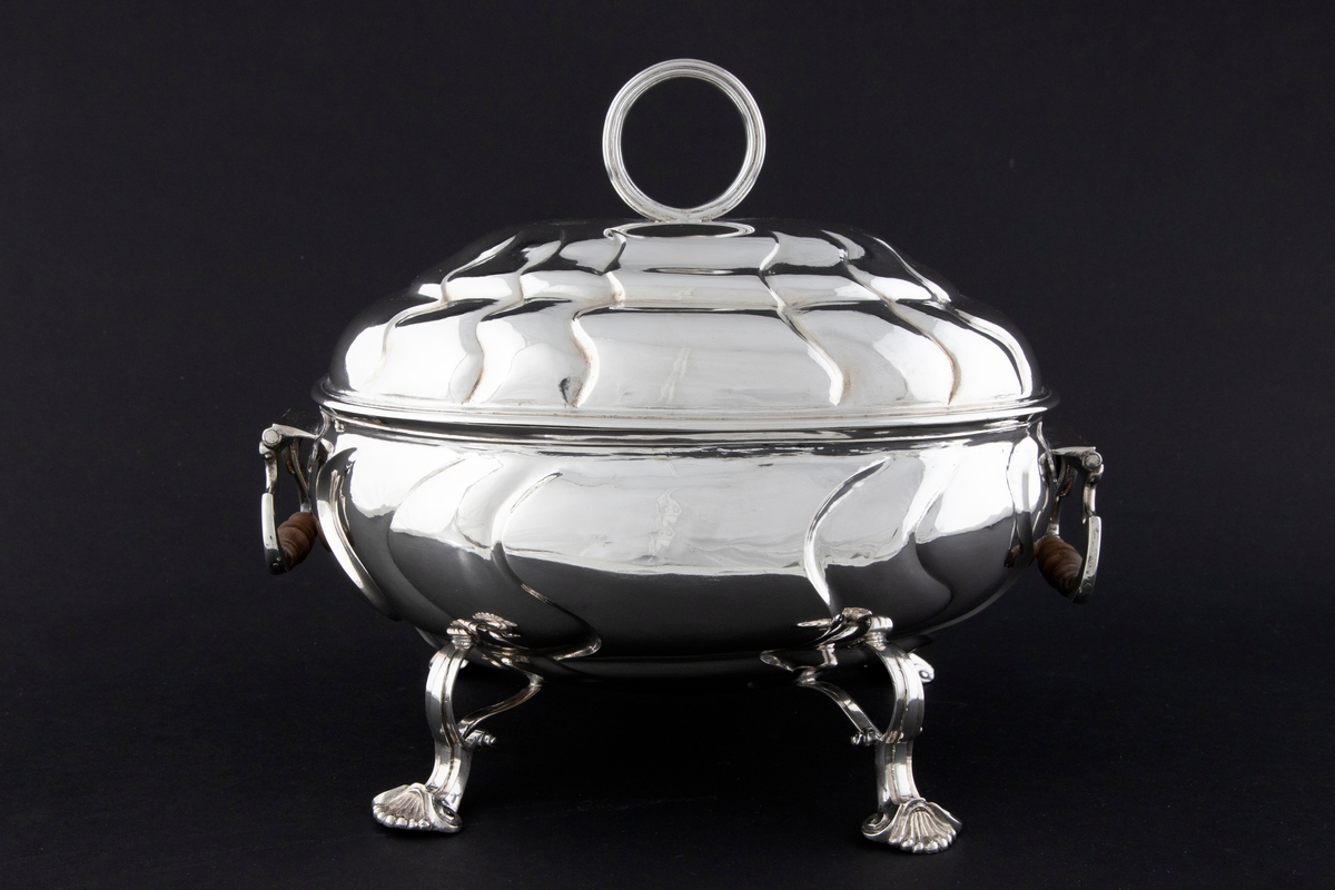 Terrin med lokk i sølv. Den har oval form og står på fire svungne bein som nederst slutter i et muslingmotiv. Korpus og det hvelvede lokket er dekorert med sveifede knekk. På hver kortside er hengslet håndtak med tre-grep. På lokket er det et ringformet grep.