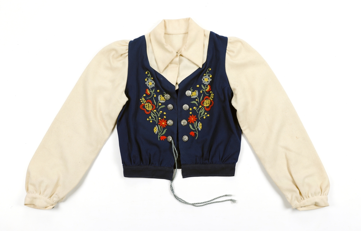 Overdel til drakt bestående av et kypertvevd blått bomullstoff brodert med ullgarn og med ermer og krage av lerretsvevd ull. Front og ermer har tinnknapper med støpt mønster. Det er hekter og trykknapper til å lukke med.
Buksa til drakta er av typen strekkbukse med press og strikk under beina. Den har glidelåslukning i siden og to lommer med glidelås i front.