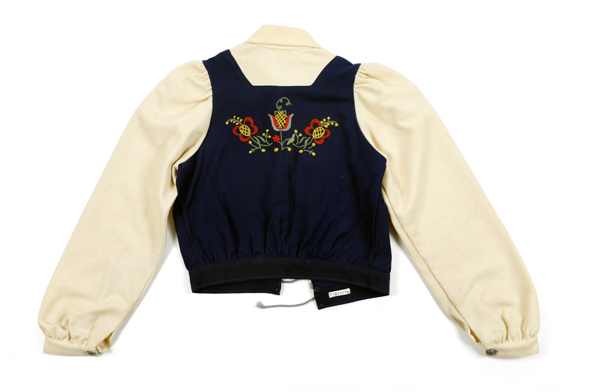 Overdel til drakt bestående av et kypertvevd blått bomullstoff brodert med ullgarn og med ermer og krage av lerretsvevd ull. Front og ermer har tinnknapper med støpt mønster. Det er hekter og trykknapper til å lukke med.
Buksa til drakta er av typen strekkbukse med press og strikk under beina. Den har glidelåslukning i siden og to lommer med glidelås i front.