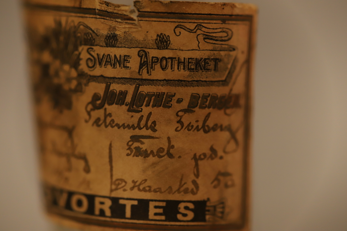 Klar glassflaske med sorte biter inni. Tynn flaske hals med papirforsegling (brutt) i rødt, med  en rød og hvit snor. Papiretikett som henger halvveis på, med Svaneapotek logo og skrift. Rødt merke under med giftvarsel.