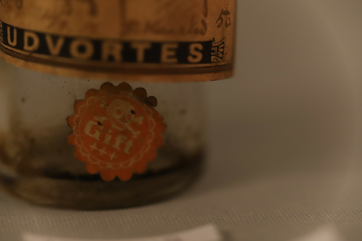 Klar glassflaske med sorte biter inni. Tynn flaske hals med papirforsegling (brutt) i rødt, med  en rød og hvit snor. Papiretikett som henger halvveis på, med Svaneapotek logo og skrift. Rødt merke under med giftvarsel.