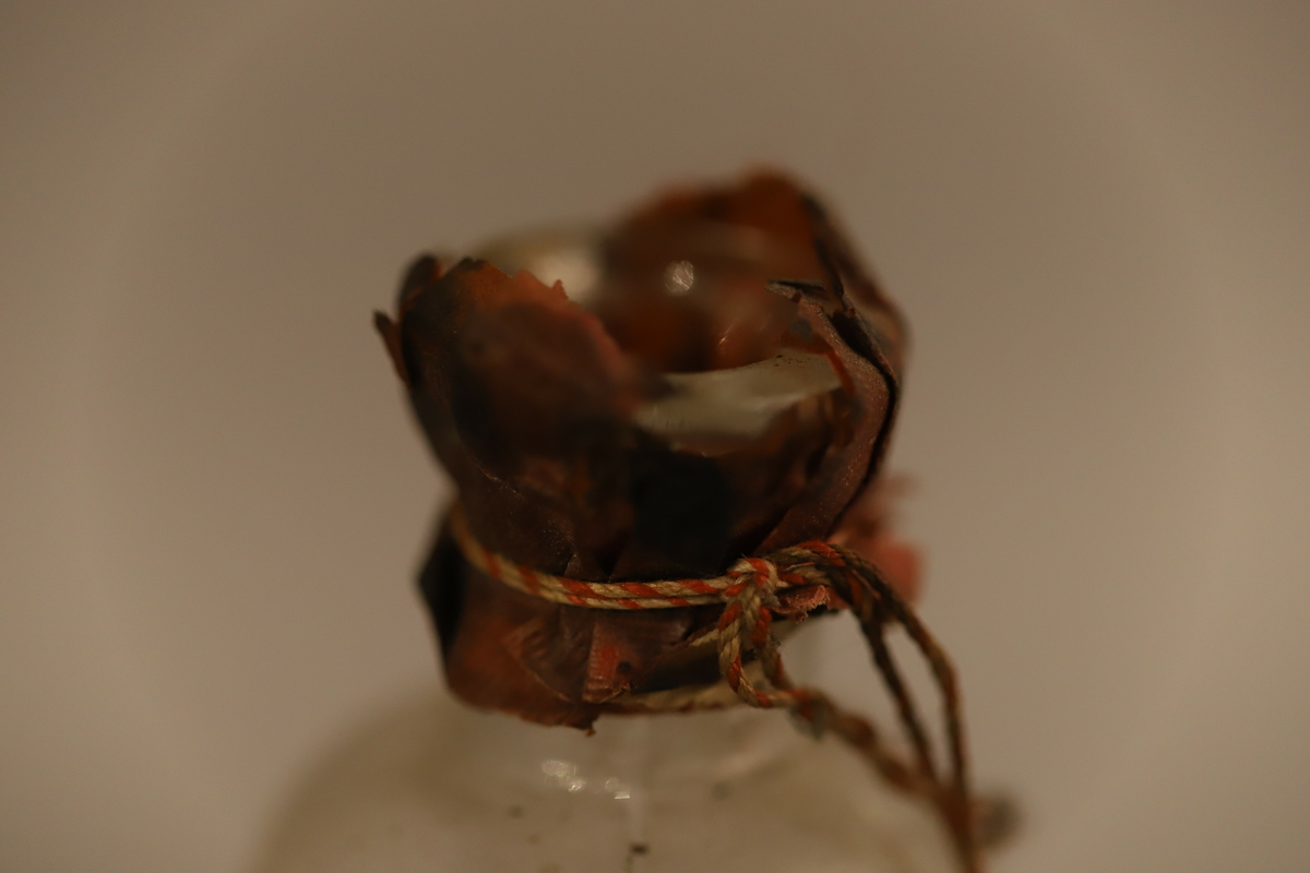 Klar glassflaske med sorte biter inni. Tynn flaske hals med papirforsegling (brutt) i rødt, med  en rød og hvit snor. Papiretikett som henger halvveis på, med Svaneapotek logo og skrift. Rødt merke under med giftvarsel.