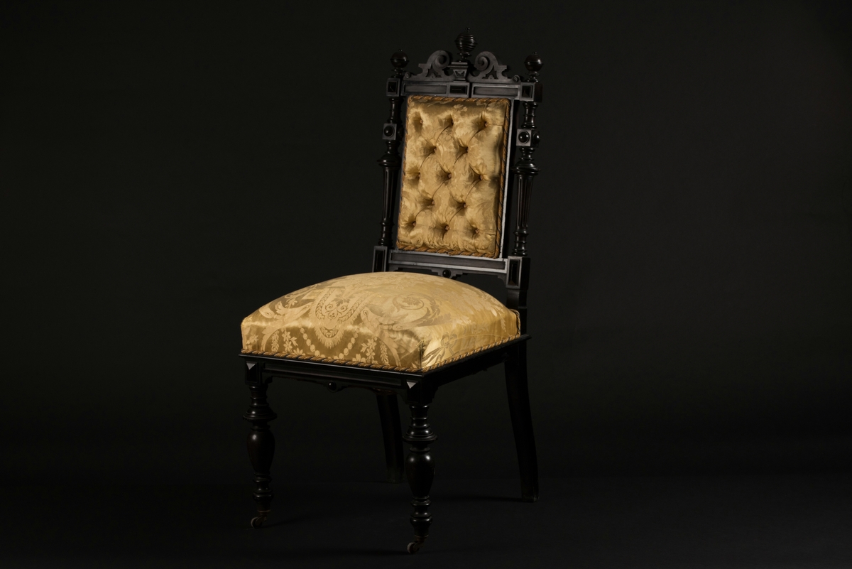 Stol i nyrenässansstil tillverkad av svart päronträ och klädd i gul sidendamast. 
Sargen och ryggstödsramen är rikt utsnidade. Det främre benparet är svarvade och försedda med trissor. Det bakre benparet är skurna, något svängda och bakåtlutande. 
Den stoppade sitsen och ryggbrickan är klädda med ett gult sidendamasttyg med invävt mönster i form av blommor, blad och band bland annat. Ryggbrickan är försedd med 13 klädda knappar.
Runt klädselns kanter mot ramverket sitter ett tvinnat kantband i gult, vitt och blått.