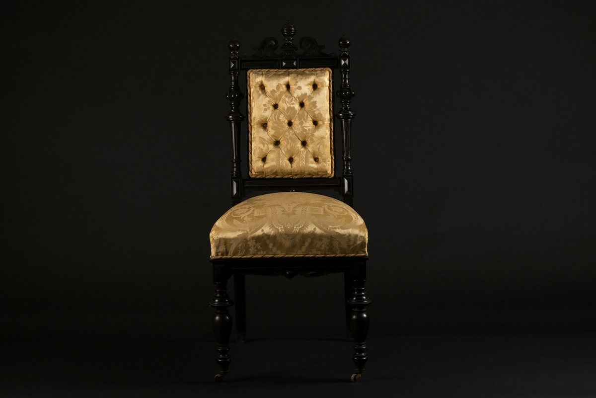 Stol i nyrenässansstil tillverkad av svart päronträ och klädd i gul sidendamast. 
Sargen och ryggstödsramen är rikt utsnidade. Det främre benparet är svarvade och försedda med trissor. Det bakre benparet är skurna, något svängda och bakåtlutande. 
Den stoppade sitsen och ryggbrickan är klädda med ett gult sidendamasttyg med invävt mönster i form av blommor, blad och band bland annat. Ryggbrickan är försedd med 13 klädda knappar.
Runt klädselns kanter mot ramverket sitter ett tvinnat kantband i gult, vitt och blått.