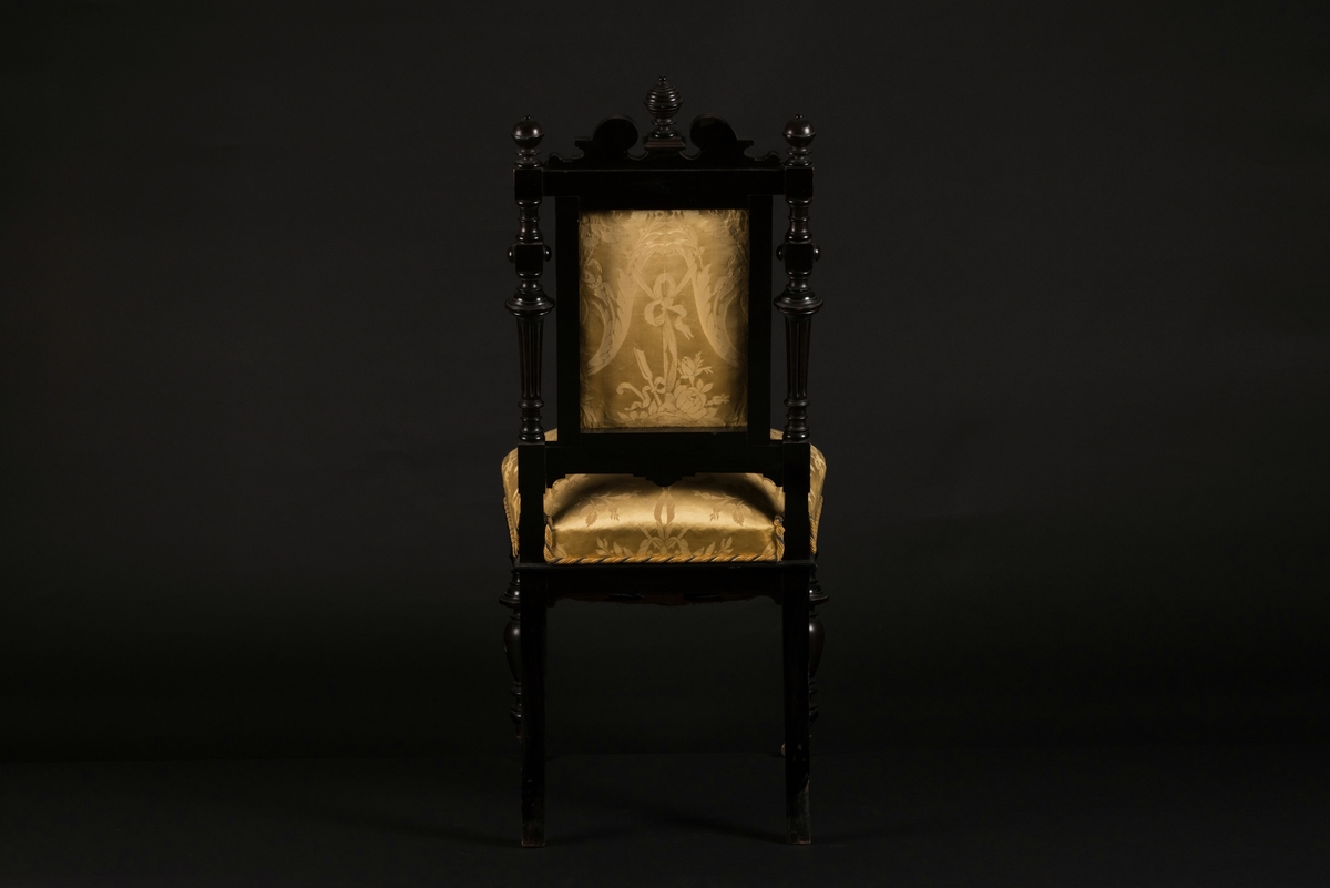 Stol i nyrenässansstil tillverkad av svart päronträ och klädd i gul sidendamast. 
Sargen och ryggstödsramen är rikt utsnidade. Det främre benparet är svarvade och försedda med trissor. Det bakre benparet är skurna, något svängda och bakåtlutande. 
Den stoppade sitsen och ryggbrickan är klädda med ett gult sidendamasttyg med invävt mönster i form av blommor, blad och band bland annat. Ryggbrickan är försedd med 13 klädda knappar.
Runt klädselns kanter mot ramverket sitter ett tvinnat kantband i gult, vitt och blått.