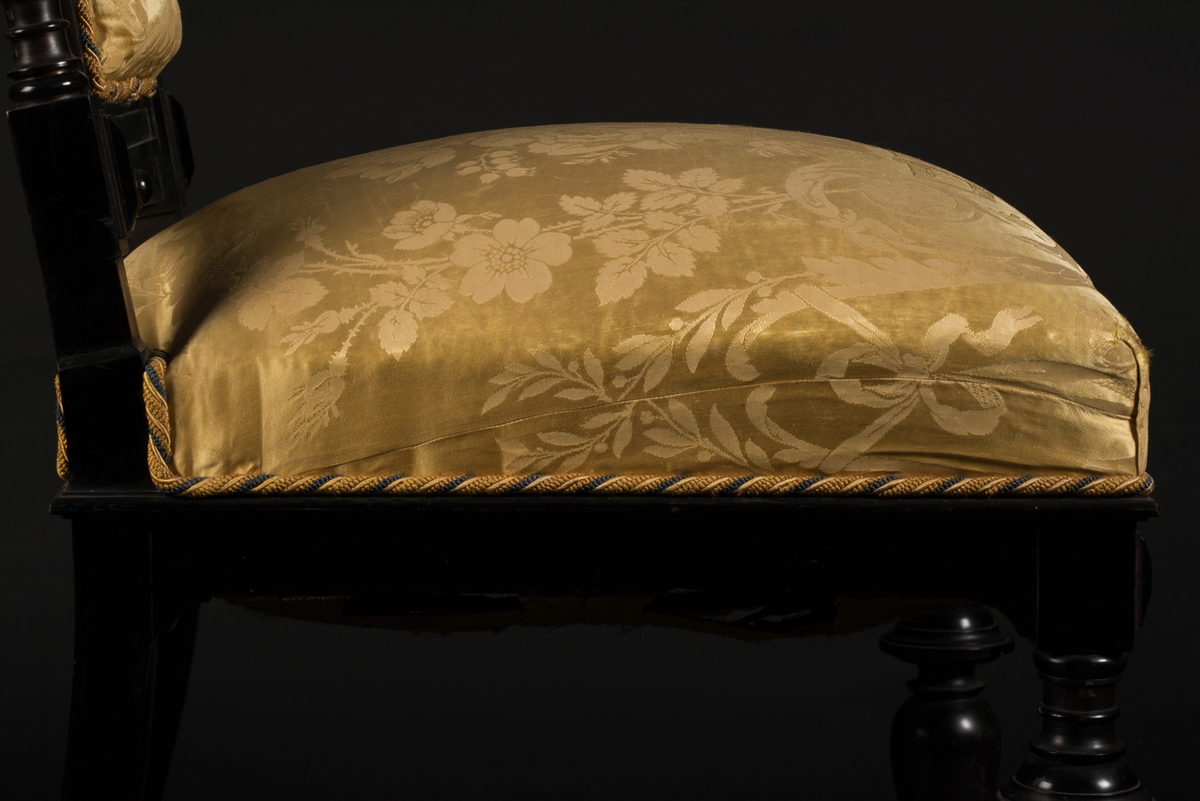 Stol i nyrenässansstil tillverkad av svart päronträ och klädd i gul sidendamast. 
Sargen och ryggstödsramen är rikt utsnidade. Det främre benparet är svarvade och försedda med trissor. Det bakre benparet är skurna, något svängda och bakåtlutande. 
Den stoppade sitsen och ryggbrickan är klädda med ett gult sidendamasttyg med invävt mönster i form av blommor, blad och band bland annat. Ryggbrickan är försedd med 13 klädda knappar.
Runt klädselns kanter mot ramverket sitter ett tvinnat kantband i gult, vitt och blått.