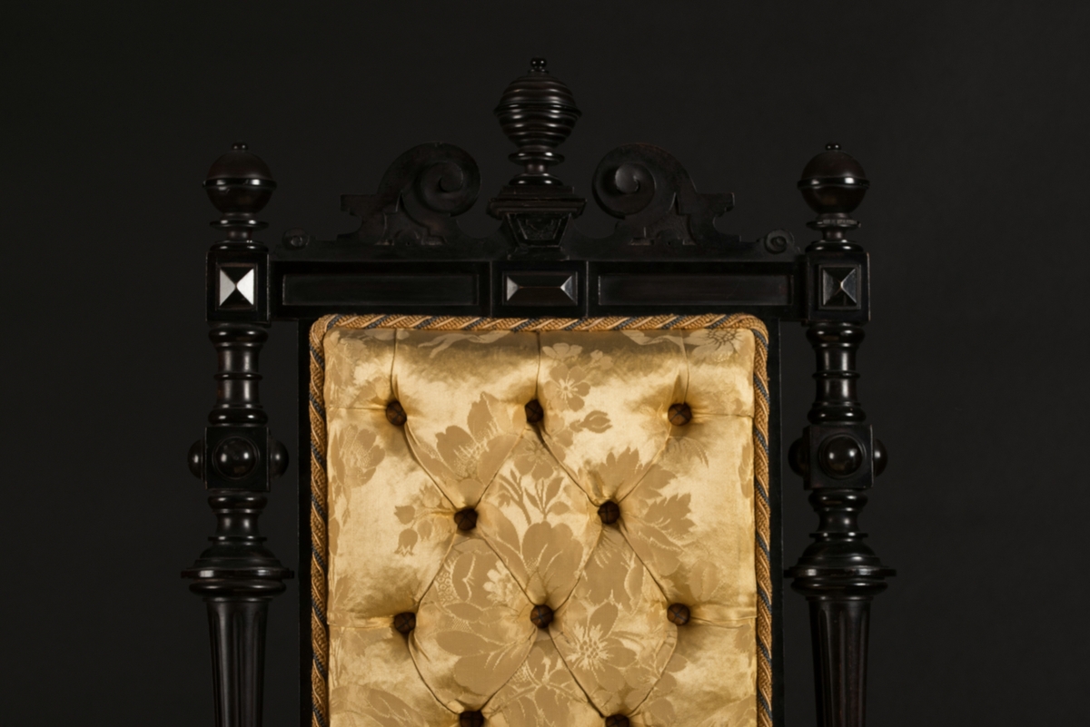 Stol i nyrenässansstil tillverkad av svart päronträ och klädd i gul sidendamast. 
Sargen och ryggstödsramen är rikt utsnidade. Det främre benparet är svarvade och försedda med trissor. Det bakre benparet är skurna, något svängda och bakåtlutande. 
Den stoppade sitsen och ryggbrickan är klädda med ett gult sidendamasttyg med invävt mönster i form av blommor, blad och band bland annat. Ryggbrickan är försedd med 13 klädda knappar.
Runt klädselns kanter mot ramverket sitter ett tvinnat kantband i gult, vitt och blått.