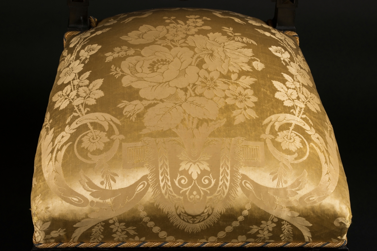 Stol i nyrenässansstil tillverkad av svart päronträ och klädd i gul sidendamast. 
Sargen och ryggstödsramen är rikt utsnidade. Det främre benparet är svarvade och försedda med trissor. Det bakre benparet är skurna, något svängda och bakåtlutande. 
Den stoppade sitsen och ryggbrickan är klädda med ett gult sidendamasttyg med invävt mönster i form av blommor, blad och band bland annat. Ryggbrickan är försedd med 13 klädda knappar.
Runt klädselns kanter mot ramverket sitter ett tvinnat kantband i gult, vitt och blått.