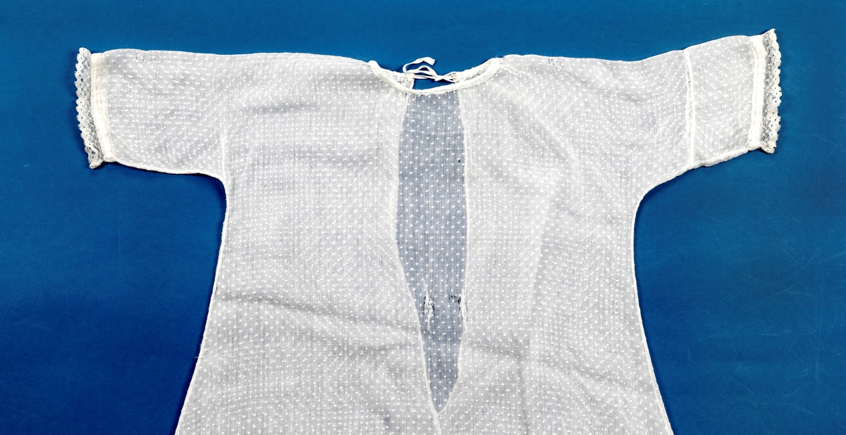 Dåpskjole med underkjole og belte.
Kjolen har rund hals med løpegang og knytesnor. Hele kjolen er klippet i ett stykke. skjøtt stoff på det ene ermet. Blondepynt i alle åpninger.  
Underkjolen er klippet i ett stykke. Rynket i halsen. Splitt bak med knepping øverst.
Bredt bånd der alle dåpsbarnas navn er brodert med henholdsvis blå og rosa skrift.