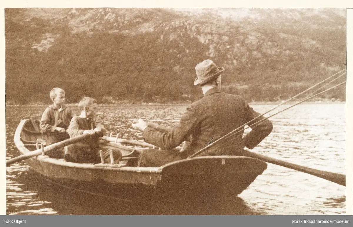 Fra Liset og Briskeroe. En mann med to gutter ror en pråm på Møsvatn. Mannen med hatt sitter bakerst i båten med ryggen til og holder i en åre. Ut stikker en fiskestang. De to guttene holder hver sin åre.