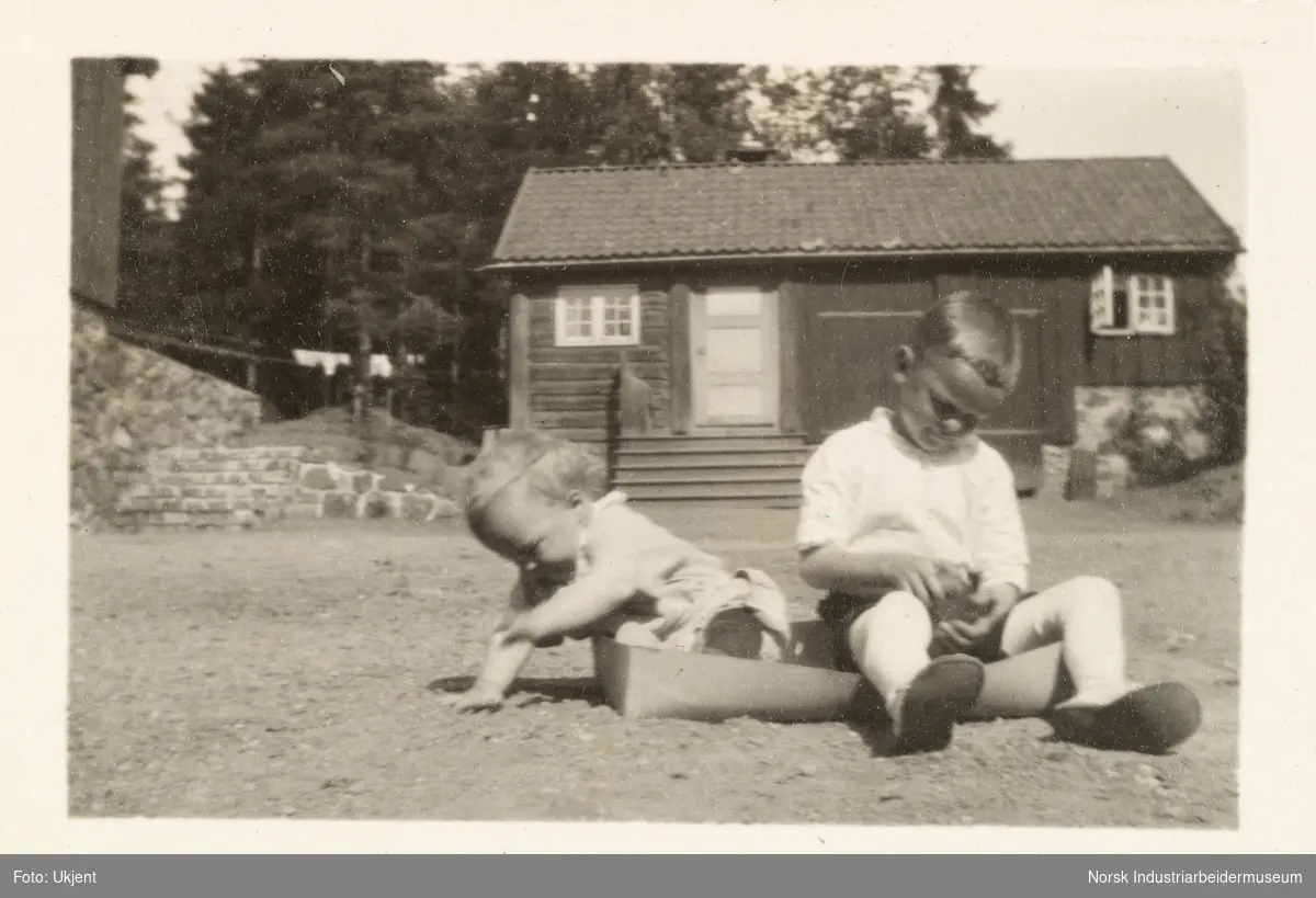 Sven Hartvig og Anton Søfren Poulsson som barn sitter i en eske på gårdsplassen til huset på Lysaker. Bak sees huseet "Brummeriet"