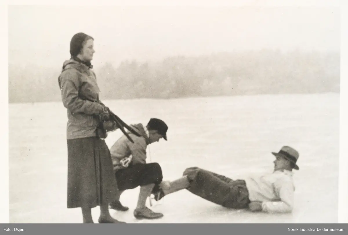 Fra Holtekilen, januar 1932. En mann ligger på isen mens en annen mann fester en skøyte på hans fot. Ved siden står en kvinne i skjørt. Alle har hodeplagg