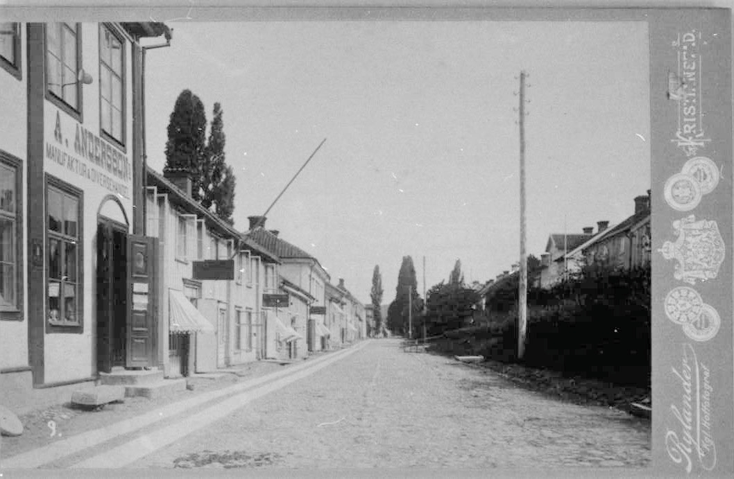 Brahegatan i Gränna mot norr. En rad butiker på västra sidan: A Anderssons Manufaktur & Diversehandel, Fröknarna Rosenborgs Sybehör, tricot & garnaffär samt E Magnussons Nya Matvaru-Handel.