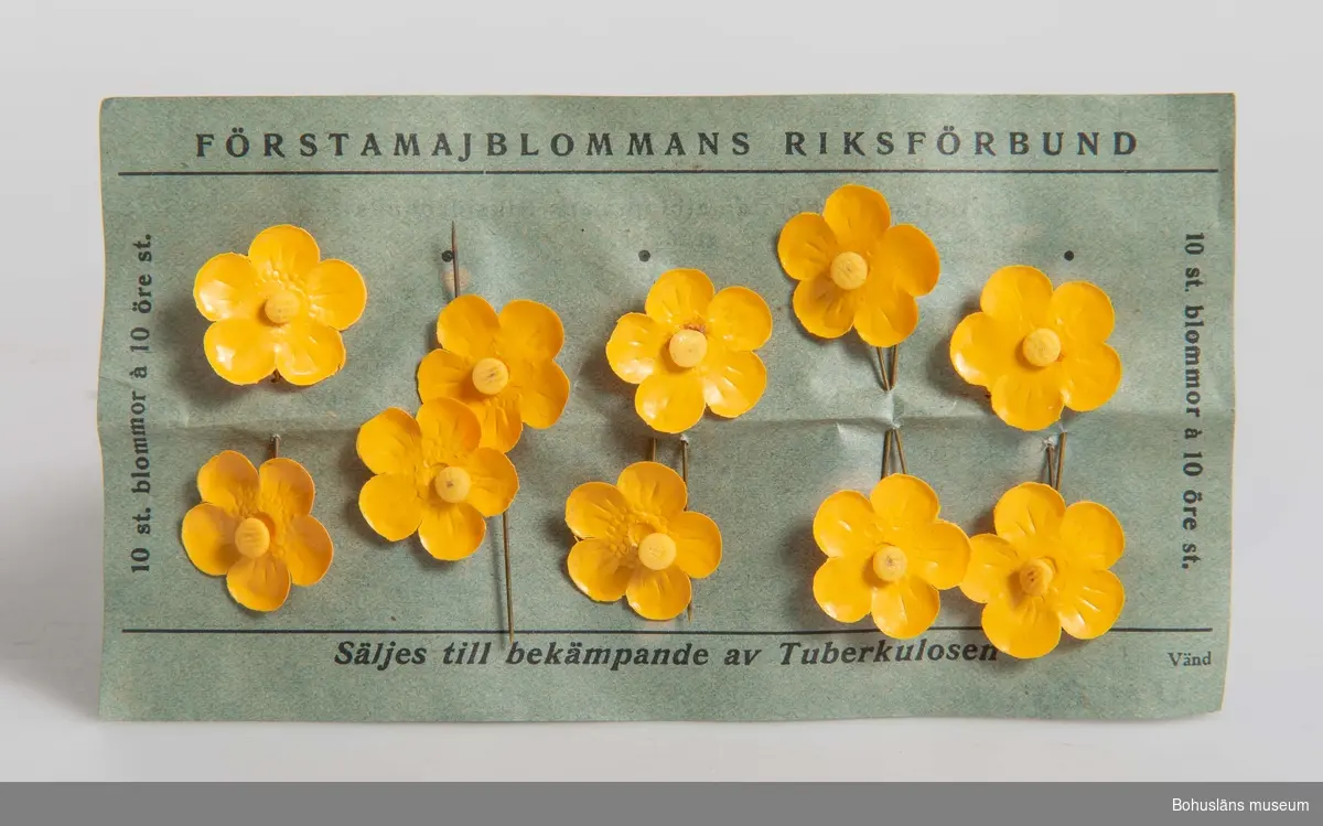 Två kartor med förstamajblommor fästade på pappersark s.k. kartor. 10 blommor per karta.
Ena karta av blågrönt papper med svart text: "Förstamajblommans Riksförbund. Säljes till bekämpande av tuberkulos. 10 st blommor a 10 öre st". På baksidan utdrag av Förstamajblommans Riksförbunds stadgar. 
På den blågröna kartan 10 gula förstamajblommor tillverkade år 1922.
Den andra kartan av vitt papper med svart text, 10 blommor från 1923. Samma text som föregående, men med tillägg "Bär ej blomman före Valborgsmässoafton".
Rektangulär grå pappask med påklistrad banderoll, som fungerat som förslutning i vitt och blått med text: "Säljes till bekämpande av tuberkulos genom Förstamajblommans Riksförbund" samt "Hugo Brusewitz Aktiebolag, Göteborg".