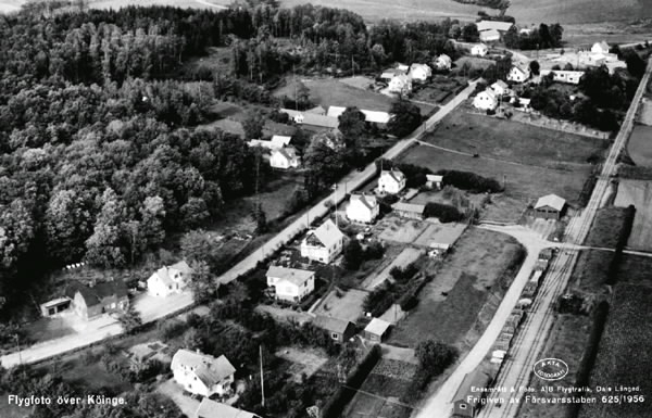 Vykort, "Flygfoto över Köinge". Enfamiljshus och byväg. Till höger ses en del av bangården till Falkenbergs Järnväg, "Pyttebanan". Lantgårdar med uthus. Odlingslandskap och skog.