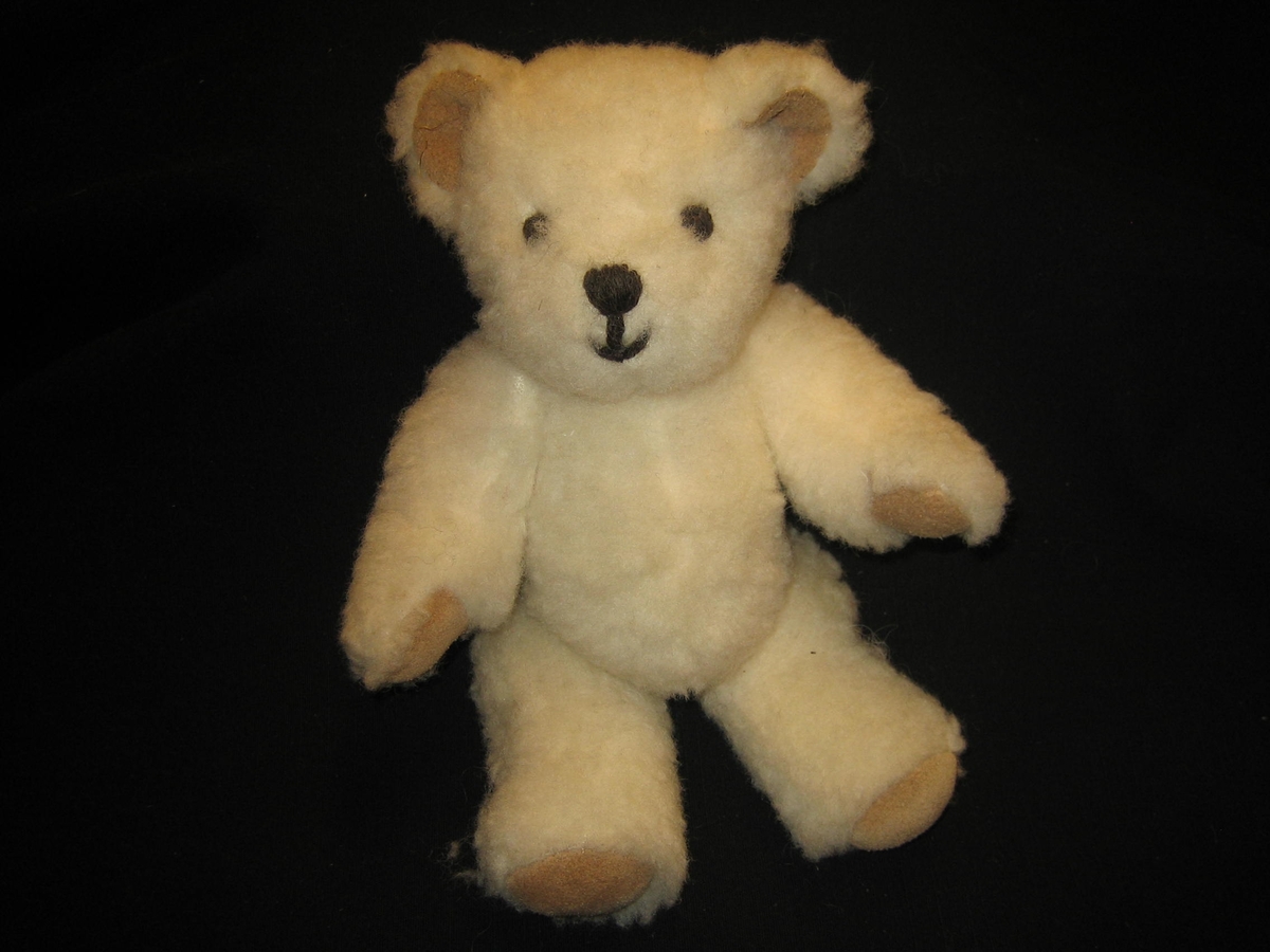 Nallebjörn i vit ullteddy med fotdynor och öron av skinn och nos och ögon är broderade med svart ullgarn. Både armar och ben är ledade. En etikett är påsydd i nacken med texten "SVENSK SLÖJD, Skaraborgs hemslöjd, SKÖVDE"