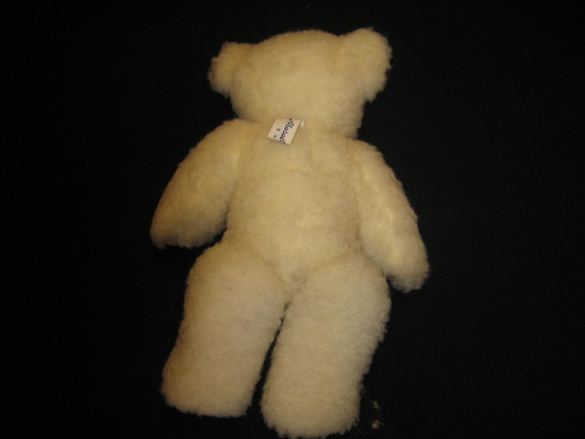 Nallebjörn i vit ullteddy med fotdynor och öron av skinn och nos och ögon är broderade med svart ullgarn. Både armar och ben är ledade. En etikett är påsydd i nacken med texten "SVENSK SLÖJD, Skaraborgs hemslöjd, SKÖVDE"