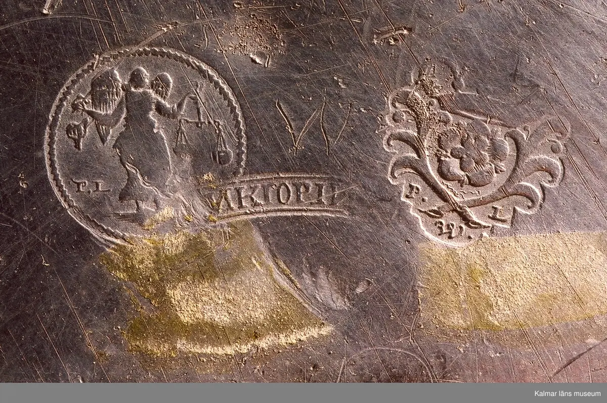KLM 5316. Tallrik av tenn. Mästarstämplar 2 små , 2 stora  med bokstäverna P.L. W, (Petter Lagerwall. Verksam under 1739-1747. Stadsstämpel: Jönkiöping och tre kronor samt bokstaven O (1755). Drevs av änkan Catharina Berg efter 25 april 1947 t.o.m. 1765. Hon är den enda tenngjutaränkan i landet som fått arbetsböcker utställda i eget namn.