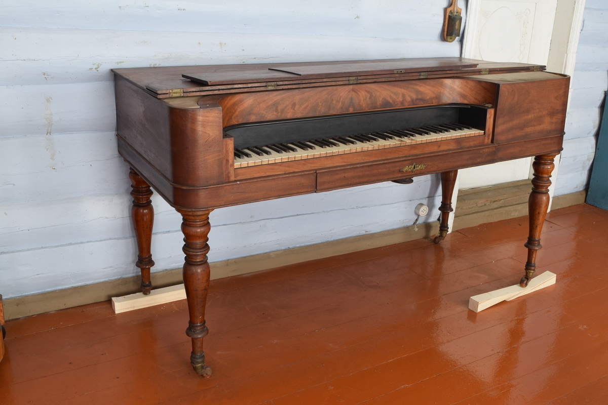 Spinett, taffelpiano i empirestil laget i mahogny. Sterkt beskadiget i strengene. Ukjent fabrikkmerke.