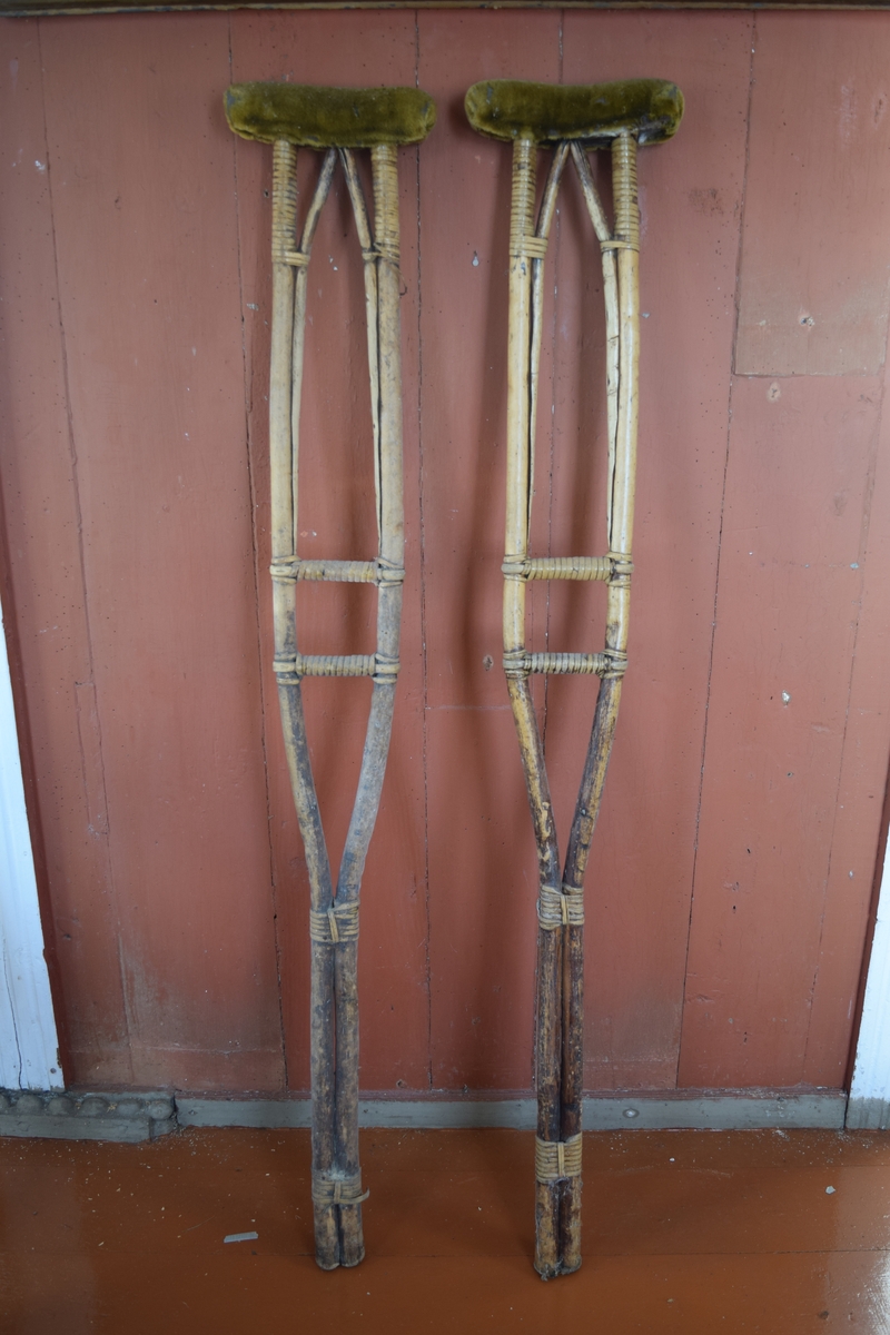 Krykker i tre med spanskrørbinding. Øverst en pute trukket i plysj. Krykkene rekker opp under armene.