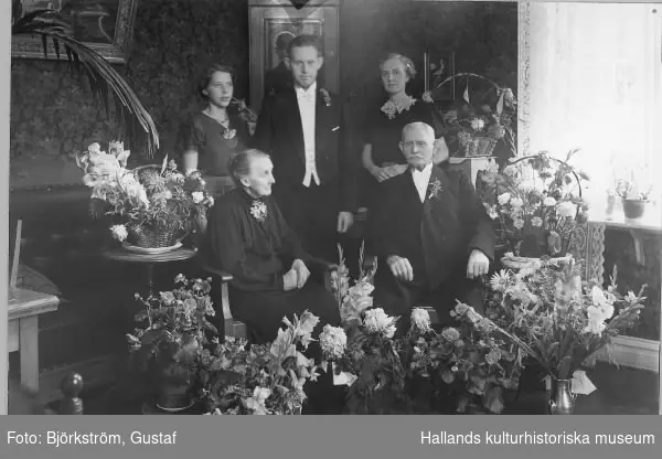 Konduktör Brudin med fru, sonen Erik, dottern Ebba och svärdotter i hemmiljö omgivna av gratulationsblomster. Troligen firas guldbröllop eller födelsedag.