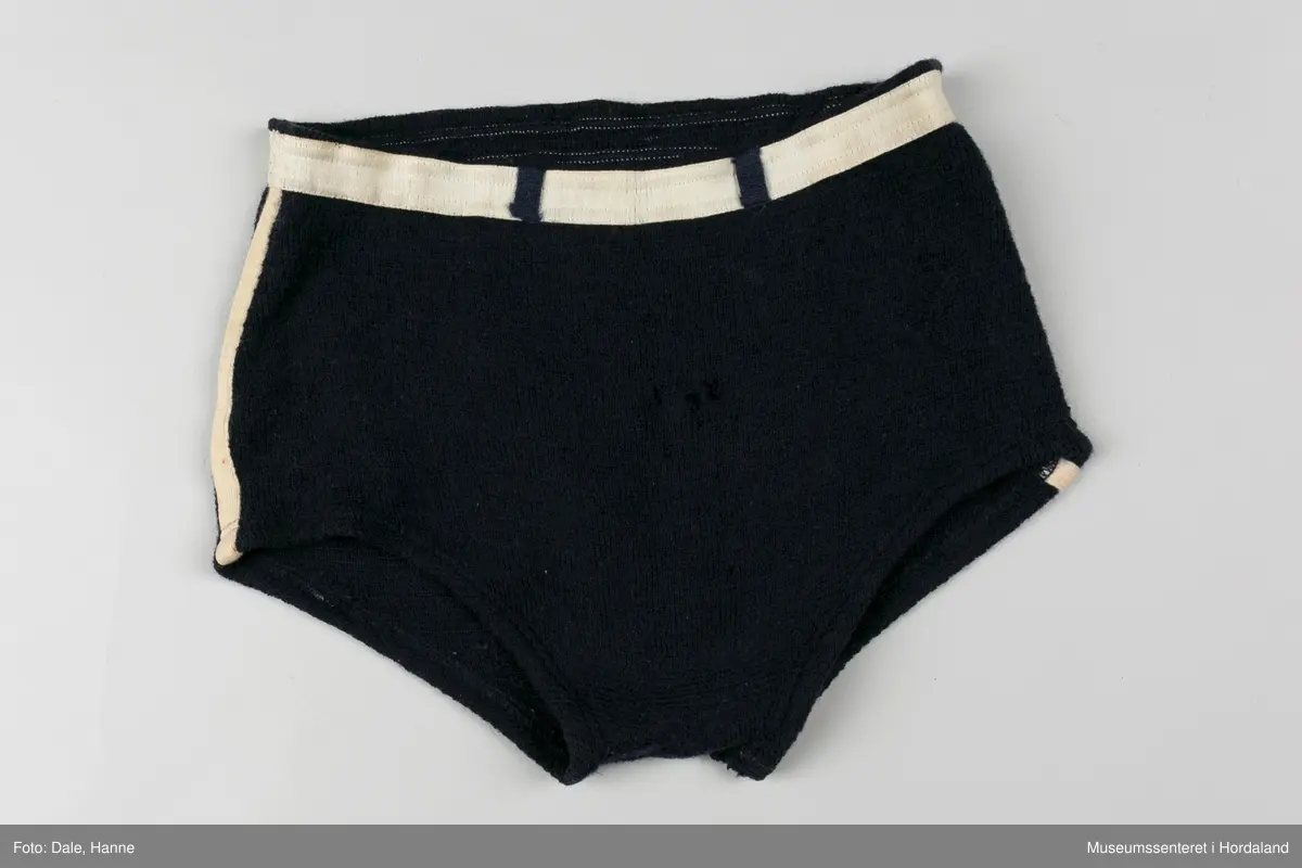 Marineblå strikka badebukse med kvit strikk i livet og kvite striper langs sidene. Str. 52, i merket Krone. Holete og godt brukt.