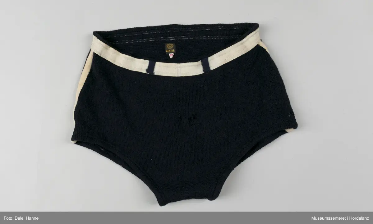 Marineblå strikka badebukse med kvit strikk i livet og kvite striper langs sidene. Str. 52, i merket Krone. Holete og godt brukt.