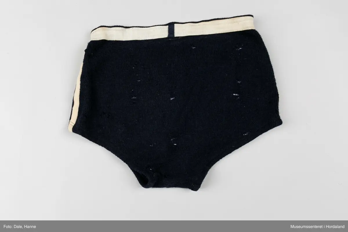 Marineblå strikka badebukse med kvit strikk i livet og kvite striper langs sidene. Str. 52, i merket Krone. Holete og godt brukt.