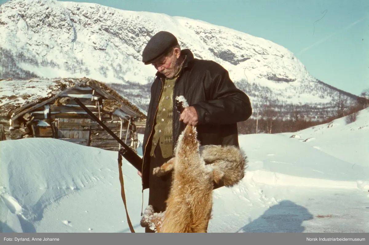 Jon Mogen i fjellet med gevær i den ene handa og en rev i den andre. I bakgrunn laftet hus og fjell dekket av snø.