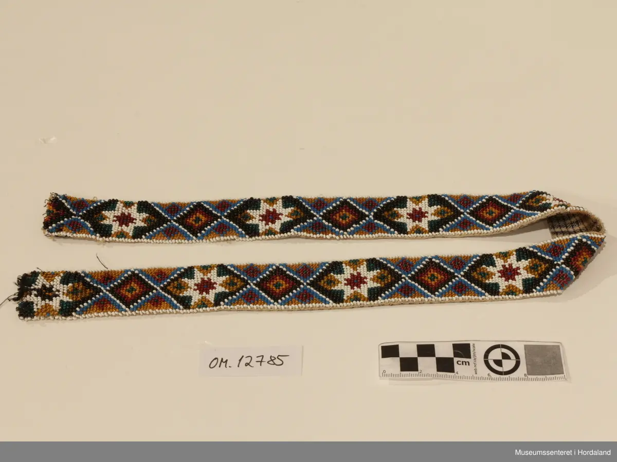 Perlebelte sydd på stramei. Mønsteret er 8-bladroser og firkanter. Beltet er fõra med eit bomullstoff. I opningen er det to par kroker og maljer (hemper).