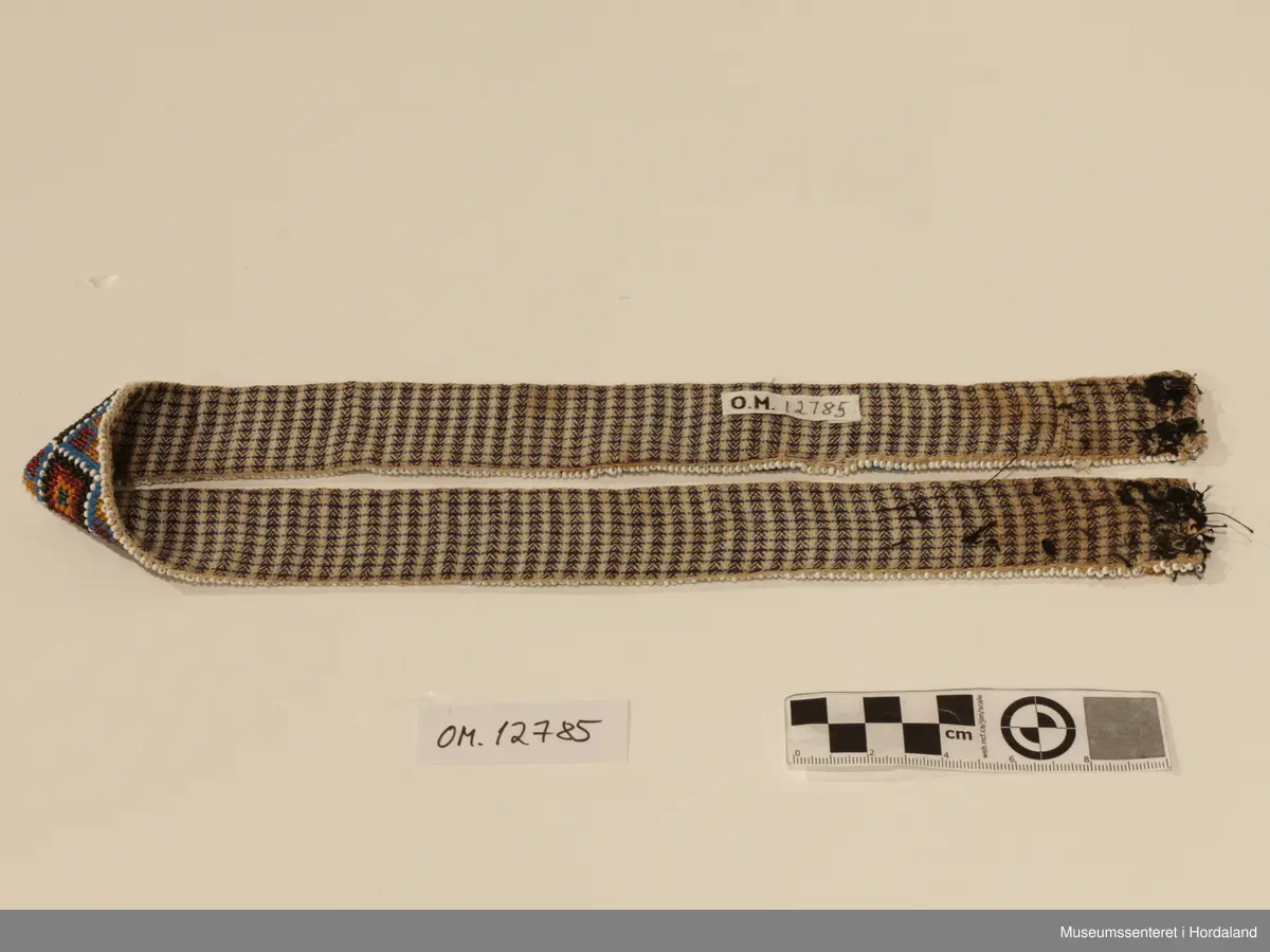 Perlebelte sydd på stramei. Mønsteret er 8-bladroser og firkanter. Beltet er fõra med eit bomullstoff. I opningen er det to par kroker og maljer (hemper).