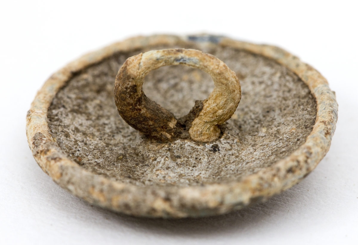 Uniformsknapp, en 20 mm bred metallknapp i militærgrønn/olivengrønn med enkel hempe bak. Rund og nokså flat form med ruglete malt overflate. Antagelig malt sink. 