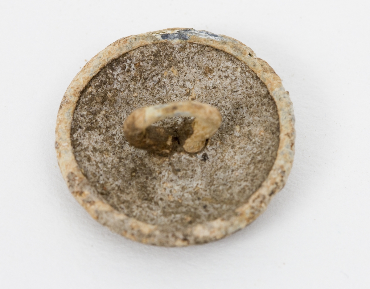 Uniformsknapp, en 20 mm bred metallknapp i militærgrønn/olivengrønn med enkel hempe bak. Rund og nokså flat form med ruglete malt overflate. Antagelig malt sink. 