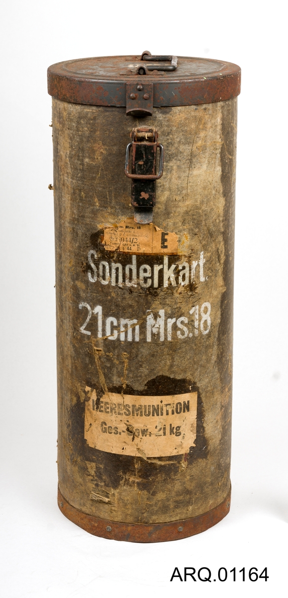 Oppbevaringsboks for ammunisjon til den største ladningen for en 21 cm Morser 18. Boksen er en sylinder og har jernlokk og jernbunn. Selve boksen er laget av meget tykk papp. Jernlokket er hengslet på med nagler inn i pappen og jernbunnen er også naglet inn i pappen. Meget solid. Lokket er foret med tekstil.