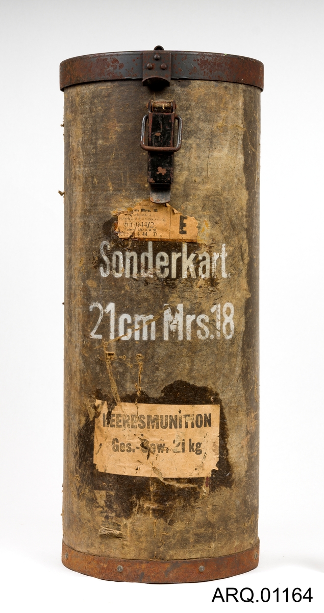Oppbevaringsboks for ammunisjon til den største ladningen for en 21 cm Morser 18. Boksen er en sylinder og har jernlokk og jernbunn. Selve boksen er laget av meget tykk papp. Jernlokket er hengslet på med nagler inn i pappen og jernbunnen er også naglet inn i pappen. Meget solid. Lokket er foret med tekstil.