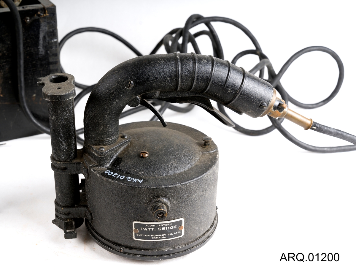 Signallampe/morselampe med batteri. Begge i svart farge. Batteriet ligger oppe i en trekasse som også er malt svart. Det hører også med en løs knott, som antagelig har falt av batteriet.
Det er en lampe av type Aldis Lantern produsert i Canada. Ved trykk på håndtaket bever speilet inne i lampen seg, med strøm og lys i lyspæren kan man da sende "SOS" ved nødsituasjoner. Lampen har et sikte på toppen.
Selve batteriet er tungt og består av 6 elementer satt sammen med ulike koblinger.