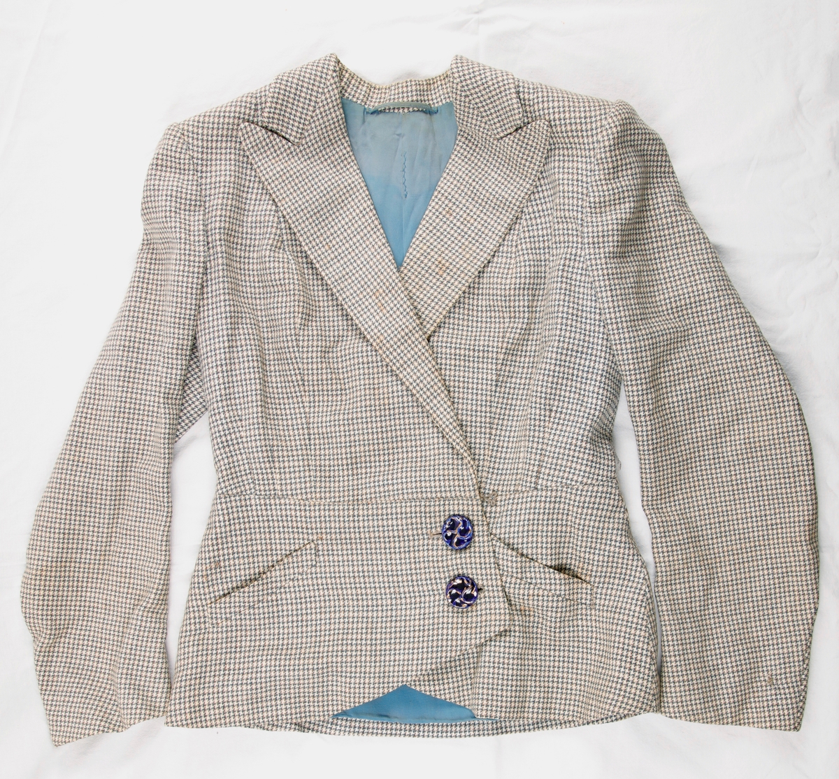 Jakke til drakt. Mønstervevet ulltekstil, foret med blått bomullstekstil. Skulderputer, jakkeslag. Lukking mot venstre, to blå knapper. To symmetrisk plasserte lommer på fremsiden av jakken.