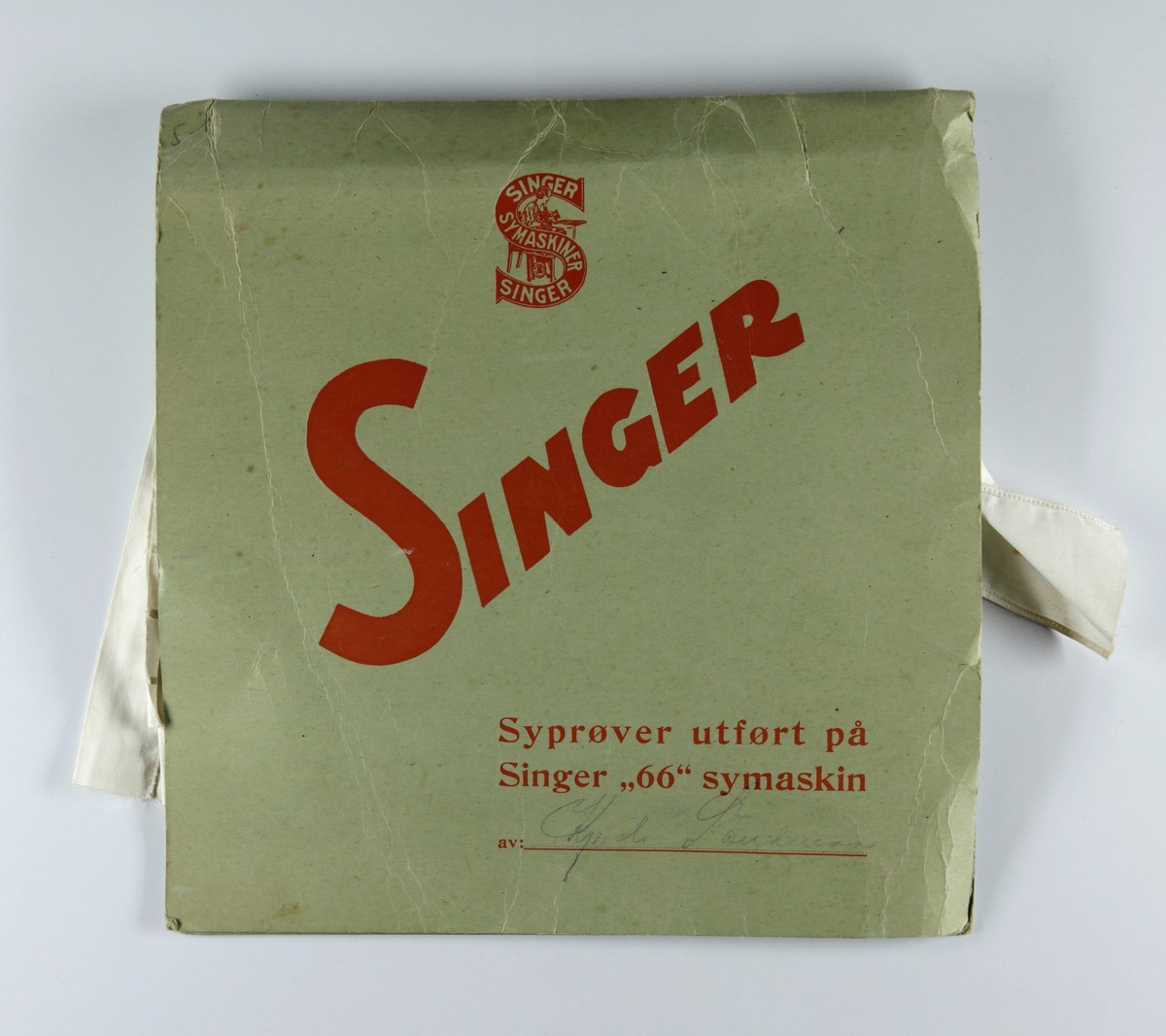 Syprøver utført på Singer "66" symaskin. Papp hefte med 13 ulike prøvelapper med ulike sømteknikker og ulike tekstilarbeid. Rødt krepp-papir rundt tekstilprøvene.