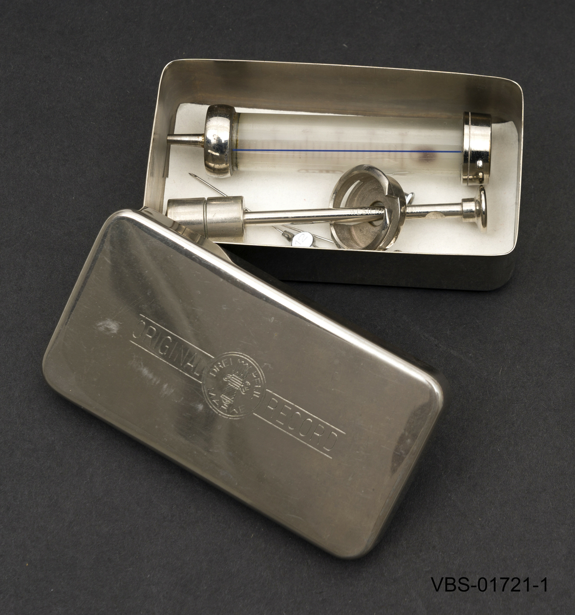 5ml sprøyte med beholder som ligger demontert i to deler, sammen med 3 sprøytespisser (nr. 12) og en stålsonde for å rengjøre nålen på innsiden.
Beholderen er en metallboks/etui i rusfritt stål med et stempel på lokket.